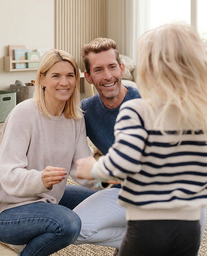 Susan und Daniel von Dein Eltern Flow sitzen lächelnd auf dem Boden im Wohnzimmer, ihre Tochter Ella steht mit dem Rücken zur Kamera vor ihnen